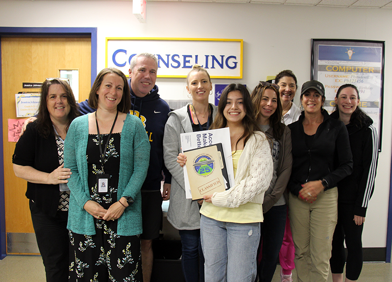 A group of eight adults stand behind a high school senior, a young woman holding a yearbook. She is wearing a white sweater. All are smiling.