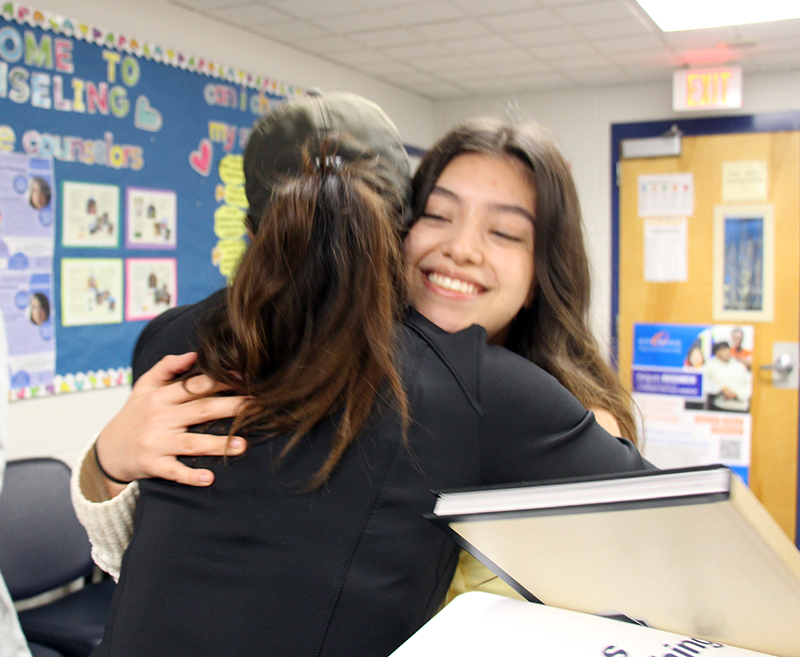 A young woman with long dark hair smiles broadly as she hugs a woman wearing black also with long black hair.