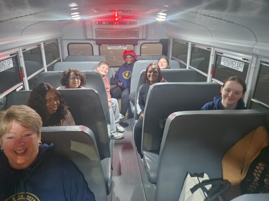 Looking down the row on a school bus. There are older kids and some adults sitting there smiling.