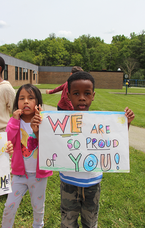 Two smaller elementary students hold a colorful sign that says We are so proud of you!