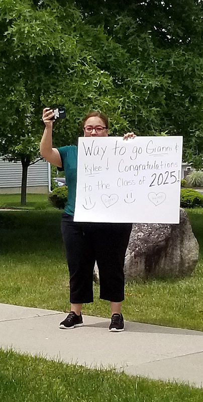 A woman stands along a street holding up a sign that says Way to go Class of 2025