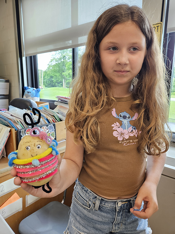 A fourth-grade girl with long blonde hair wearing a brown shirt holds a potato that is decorated with brightly colored pipe cleaners.