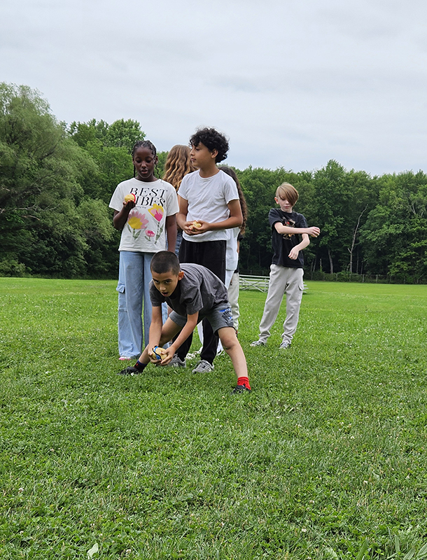 Four fourth-grade boys stand on a grass field. One is bending over with a potato in his hands.