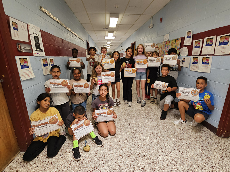 A large number of fourth-grade students stand and sit in a hallway. They are holding certificates.