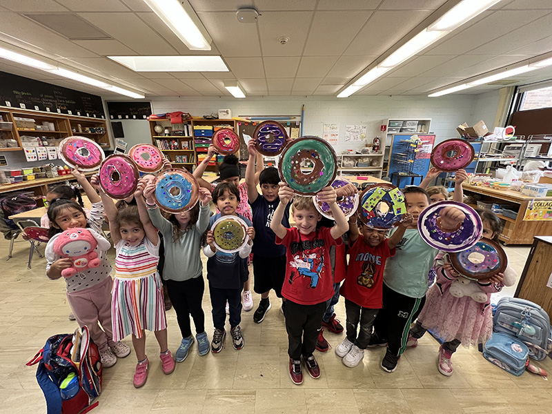 An entire class of younger elementary kids holdin gup their artwork that looks like donuts.