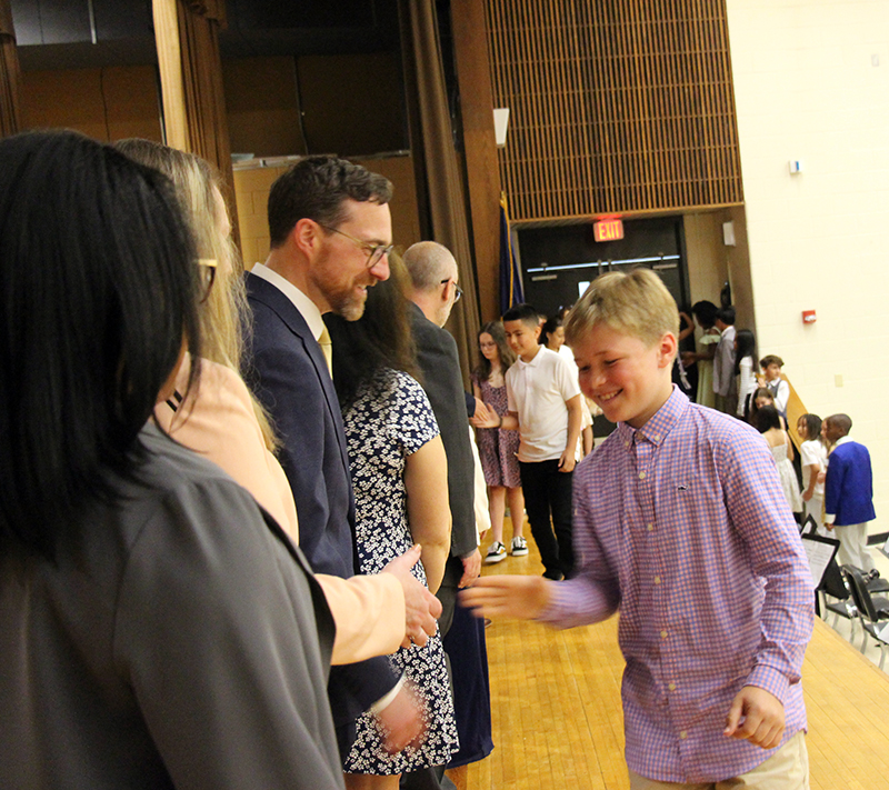 A fifth-grade boy in a button down shirt smiles as he shakes the hand of an adult on a stage.