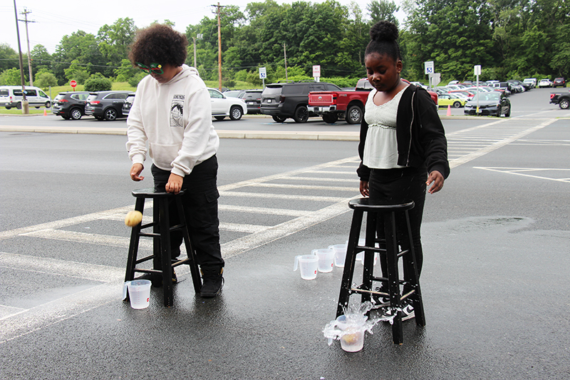 Two fourth-grade boys stand at tall stools. On the ground are two measuring cups filled with water. They push their potatoes off the stool into the cups. One potato is halfway there and another made a big splash.