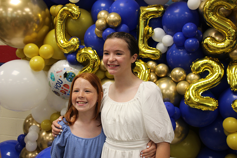 A background of blue and gold balloons. Two girls in pretty dresses stand in front and smile.