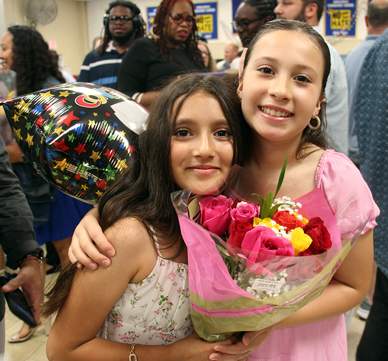 Two fifth-grade girls hug each other while holding a bouquet of flowers.