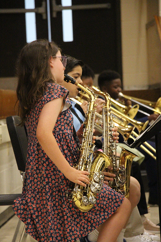 A fifth grade girl plays a saxophone. There are other musicians behind her.