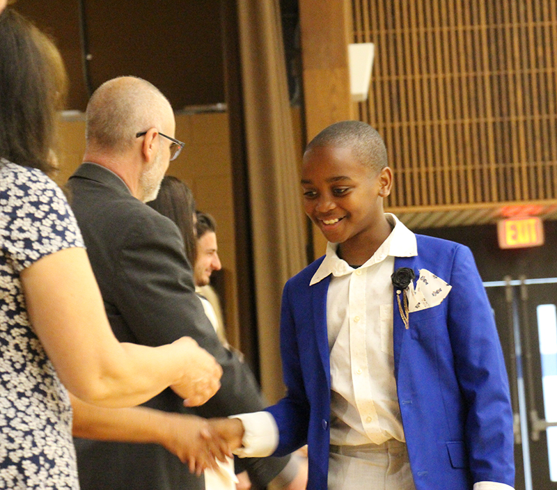 A fifth grade kid in a white shirt and blue jacket smiles and shakes the hand of someone in a line.