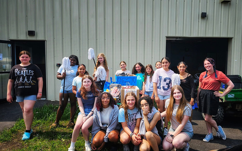 A group of 14 middle school students and an adult hold or surround a large amount of  that are being donated.
