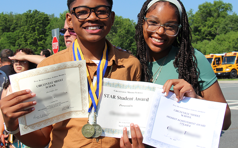 An eighth-grade boy smiles and holds three certificates. He is standing with a young woman who is also smiling.