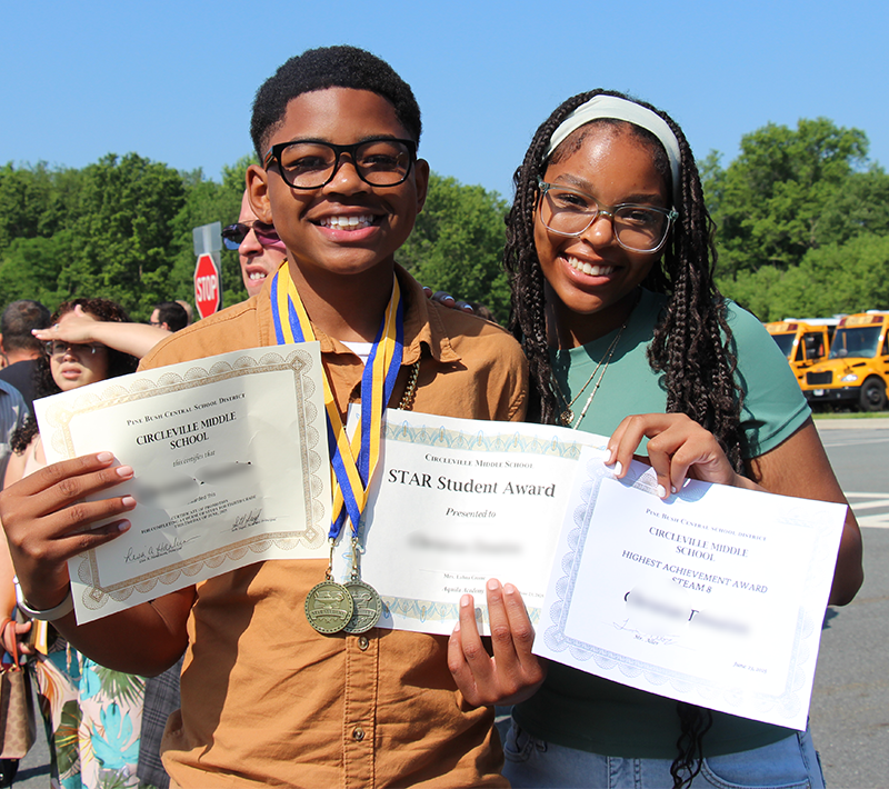 An eighth-grade boy smiles and holds three certificates. He is standing with a young woman who is also smiling.