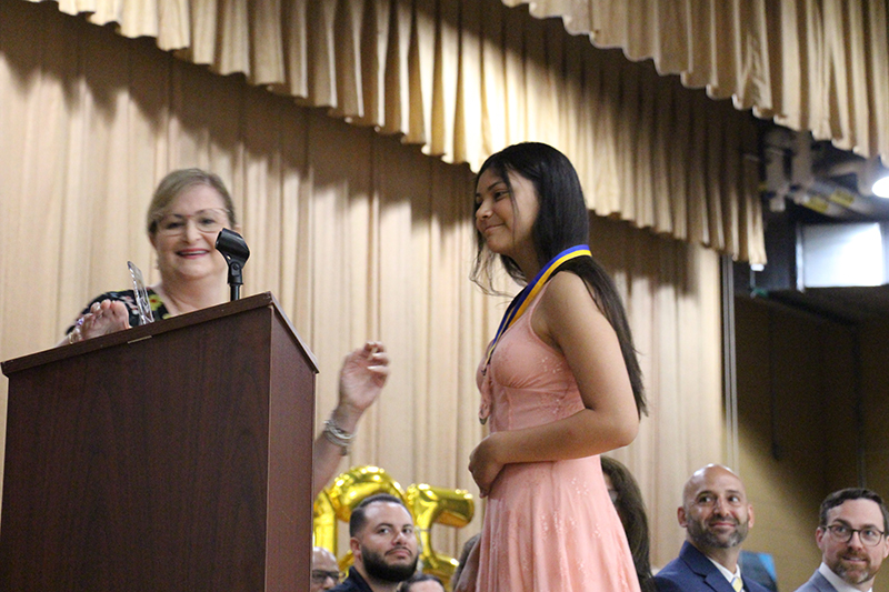 A young woman in a peach colored dress has a metal around her neck and is smiling with a woman standing nearby.