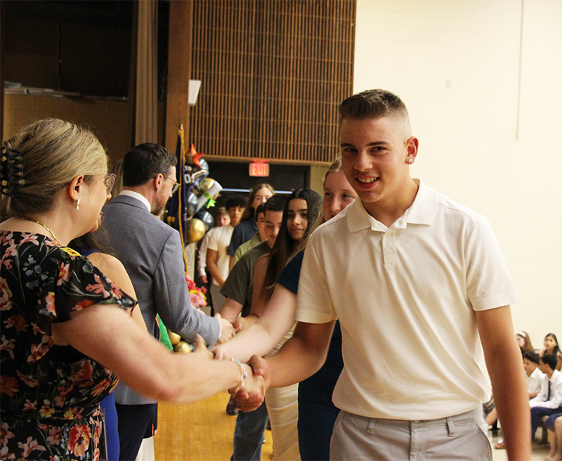 A young man shakes hands with a row of adults. He is smiling.