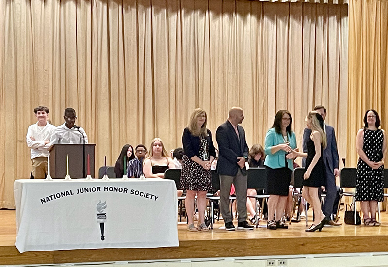 Two young people stand at a podium speaking. A line of adults are to the right shaking hands with a middle-school age young woman.
