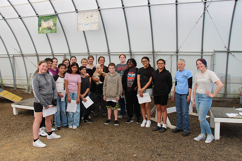 A group of 15 middle school kids stand with three adults, one of whom is holding a little white dog. They are all smiling.
