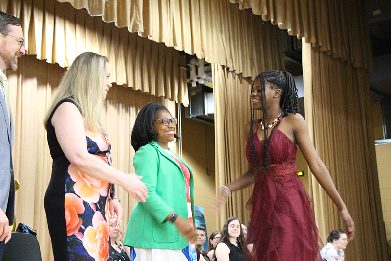 A young woman in a maroon dress shakes hands with a woman on a stage. Another woman is next to her and waiting to shake hands.