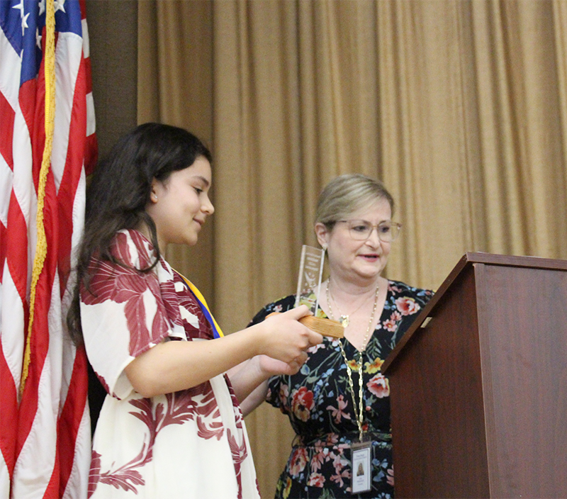An eighth-grade girl holds a small plaque and stands next to a woman in a flowered dress.