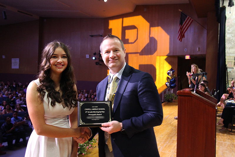 A man in a suit and tie hands a plaque to a young woman with long dark hair, wearing a white dress. Behind them on the wall is a large PB.