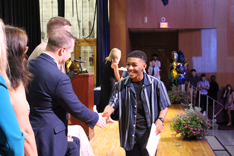 A smiling young man shakes hands with a man in a suite on a stage.