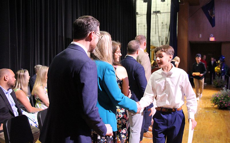 A n aighth-grade boy in a white button down shirt and blue pants shakes hands with a woman in a blue blazer. They are on a stage with other adults around them.