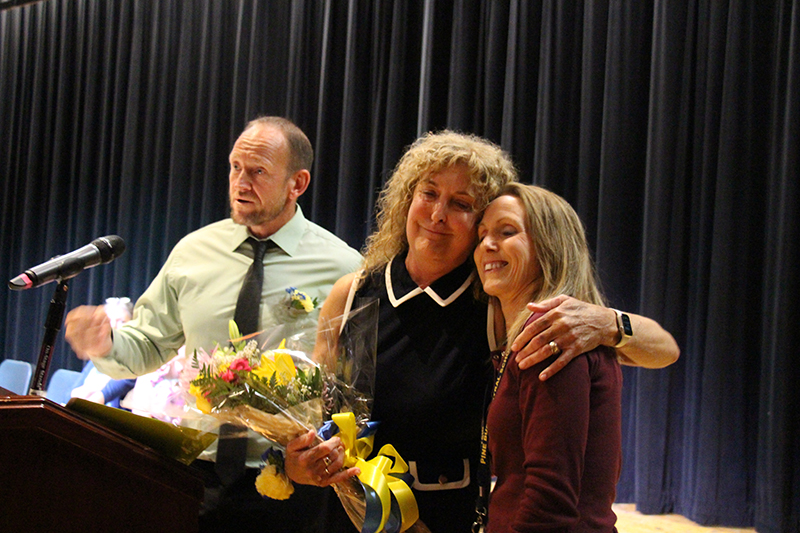 A woman with long blonde hair holds flowers and hugs a woman next to her. They are smiling. A man is in the background.