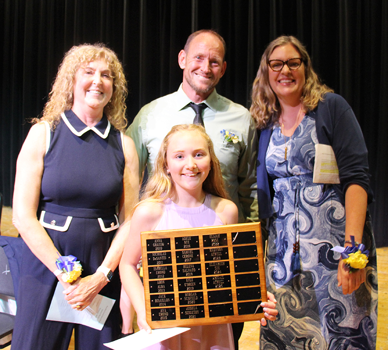 A girl with long blonde hair stands in front of three adul5ts - a man in the center and two women. She is holding a plaque.