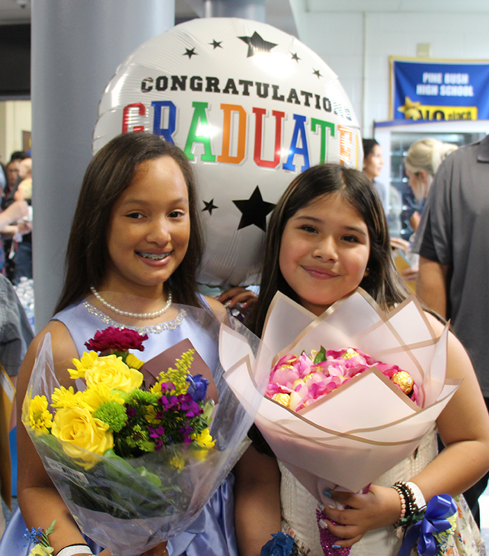 Two fifth-grade girls hold bouquets of flowers and smile. Behind them is a large balloon that says Congratulations Graduate.