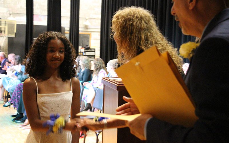A young woman in a white dress takes an envelope from a woman with long blonde hair.