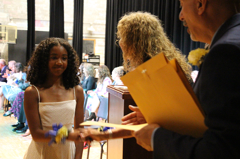 A young woman in a white dress takes an envelope from a woman with long blonde hair.