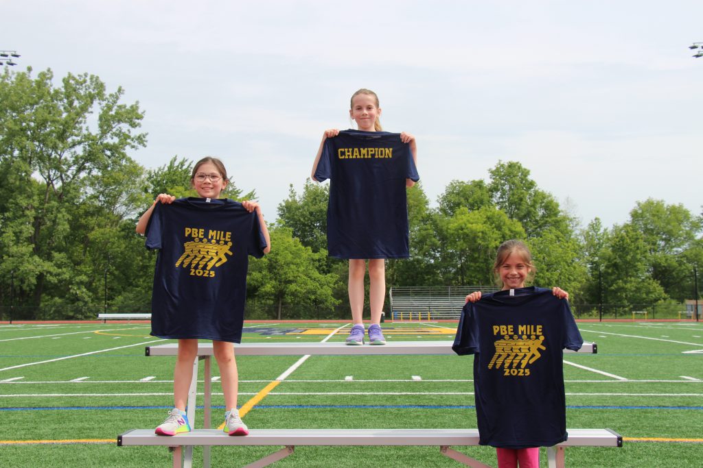 Three fourth grade girls stand in first, second and third places holding up blue tshirts.