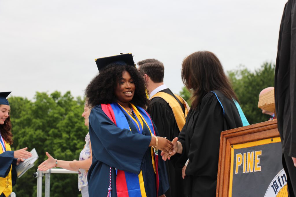A young woman wearing a blue cap and gown smiles as she shakes hands after getting her diploma.