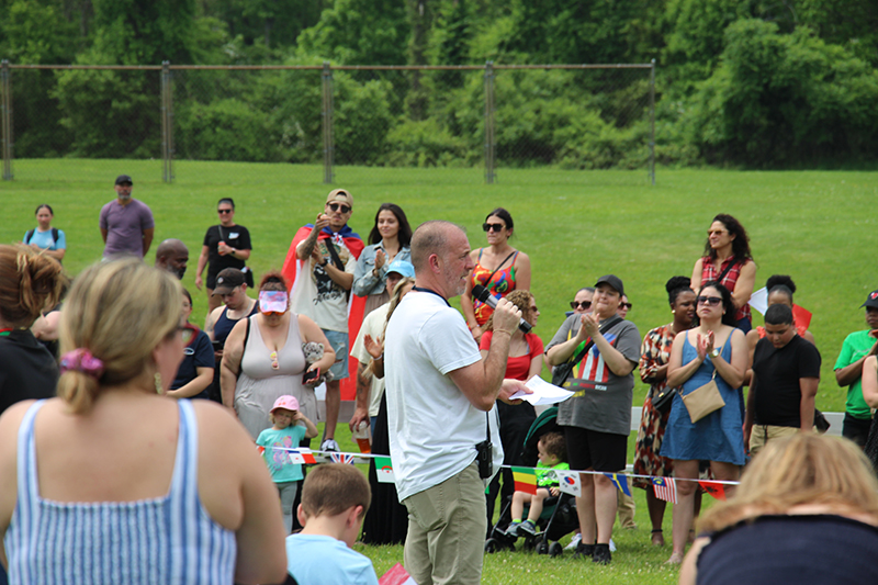 A large crowd of people stand and listen as a man talks into a microphone. They are outside on green grass.