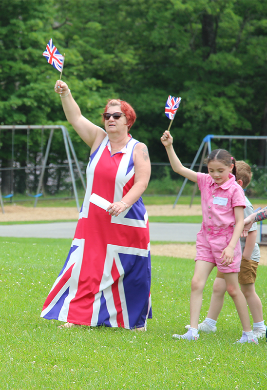 A woman in a dress like the flag of England holds up a flag. She is followed by other elementary kids holding up flags.