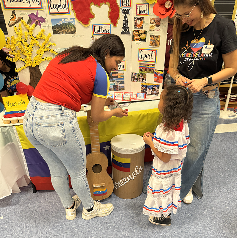 Two adults lean in to talk to a little girl at a display about a country. There is a drum in front of it.