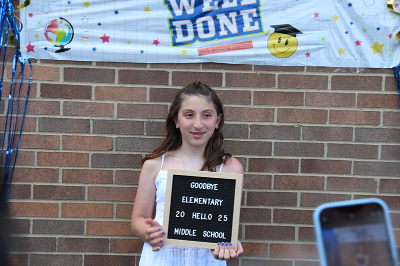 A girl in a white dress stands under a banner that says Well Done. She is holding a sign that says Goodbye Elementary Hello Middle School 2025