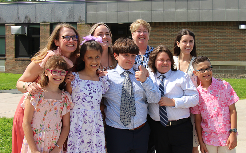 A group of five fifth-grade students - two girls and three boys - stand with four adults, their teachers.