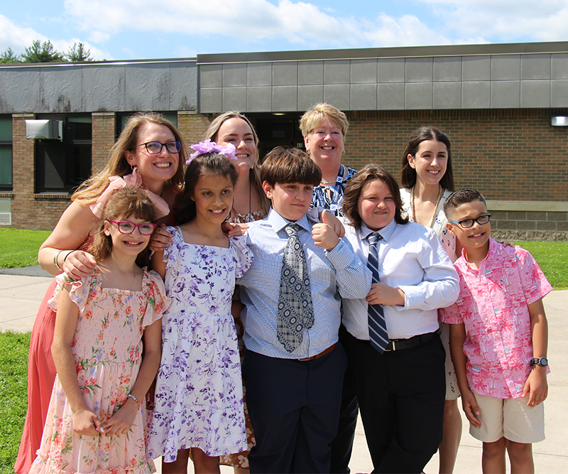 A group of five fifth-grade students - two girls and three boys - stand with four adults, their teachers.