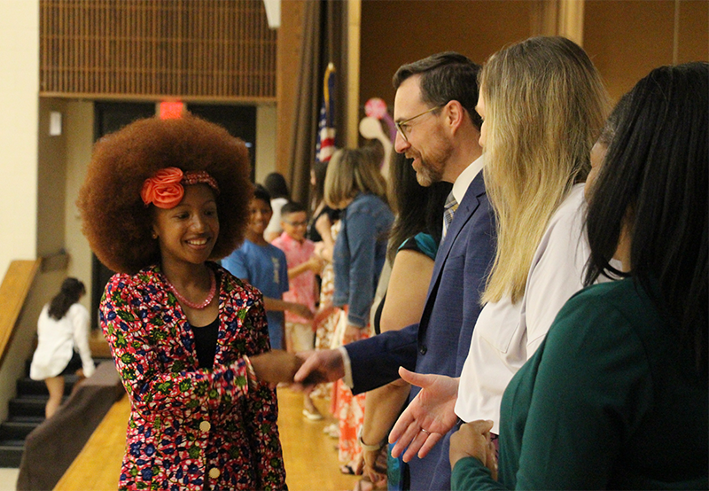 A fifth-grade girl wearing a flowered jacket  and an orange bow in her hair smiles and shakes hands with a man on a stage. There are many others waiting to shake hands.