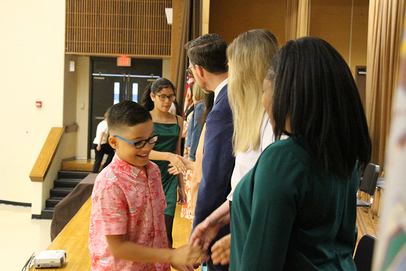 A fifth-grade boy with short dark hair and glasses shakes hands with a woman on a stage.