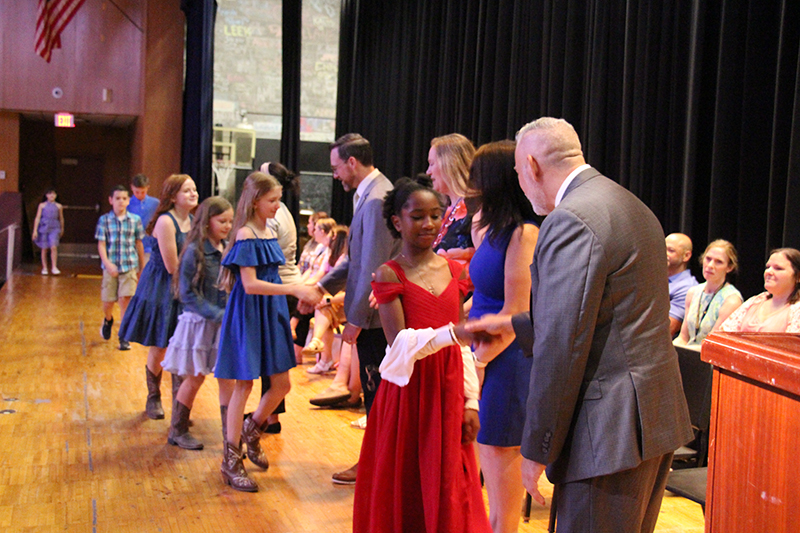 Several fifth-grade students shake hands with adults on a stage.