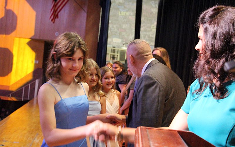A fifth-grade girl in a light blue dress shakes hands with a woman on a stage.