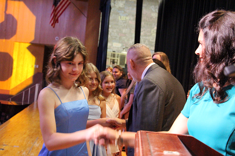 A fifth-grade girl in a light blue dress shakes hands with a woman on a stage.
