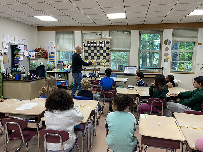 A man stands in front of a class of students with a large chess board projected. He is teaching about the game.