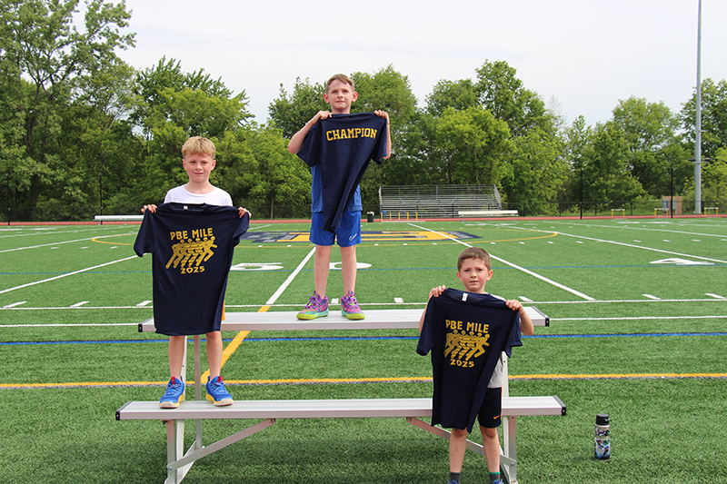 Three third grade boys stand in first, second and third places holding up blue tshirts.