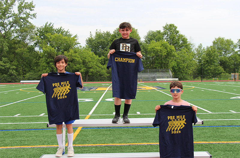 Three fourth grade boys stand in first, second and third places holding up blue tshirts.