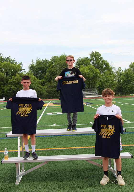 Three fifth grade boys stand in first, second and third places holding up blue tshirts.