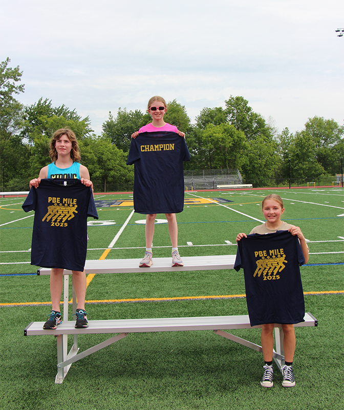 Three fifth grade girls stand in first, second and third places holding up blue tshirts.